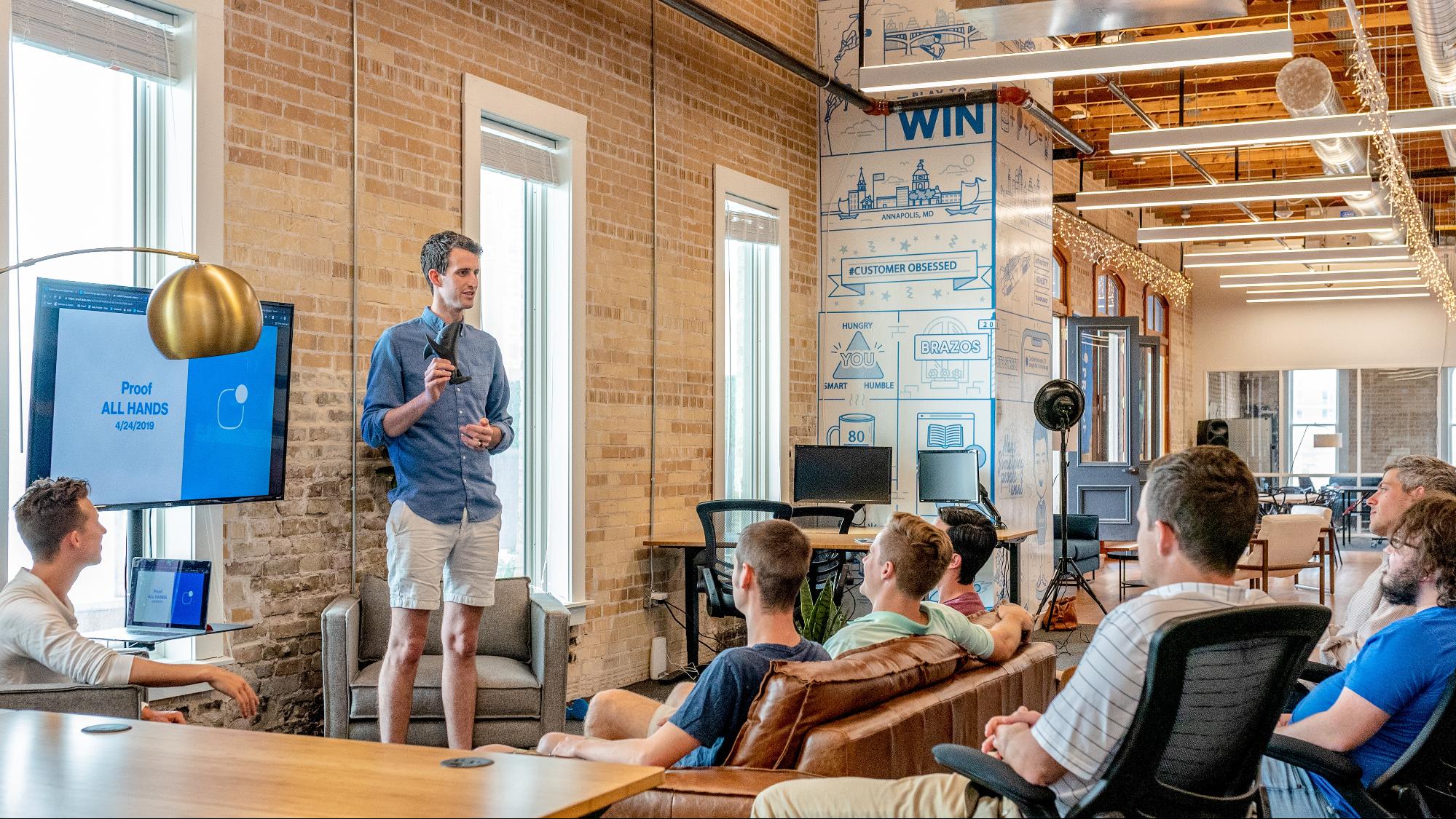 A white guy with a remote and screen standing in front of crowd.