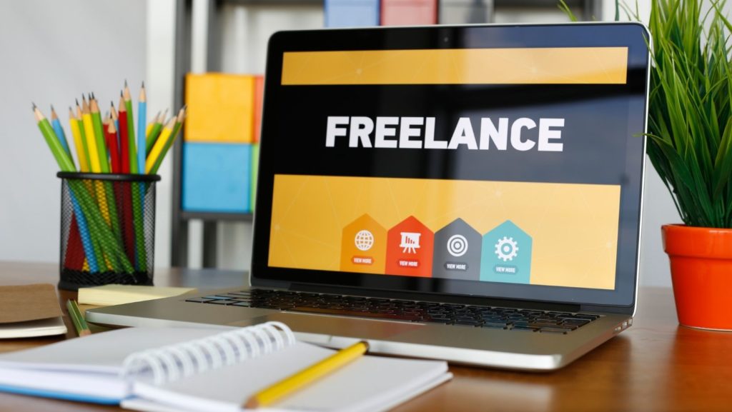Laptop with "freelance" on the display with a notebook in the foreground and pencils in the background